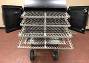 Barbecue grill with open lid and three metal trays on wheels on a red brick floor.