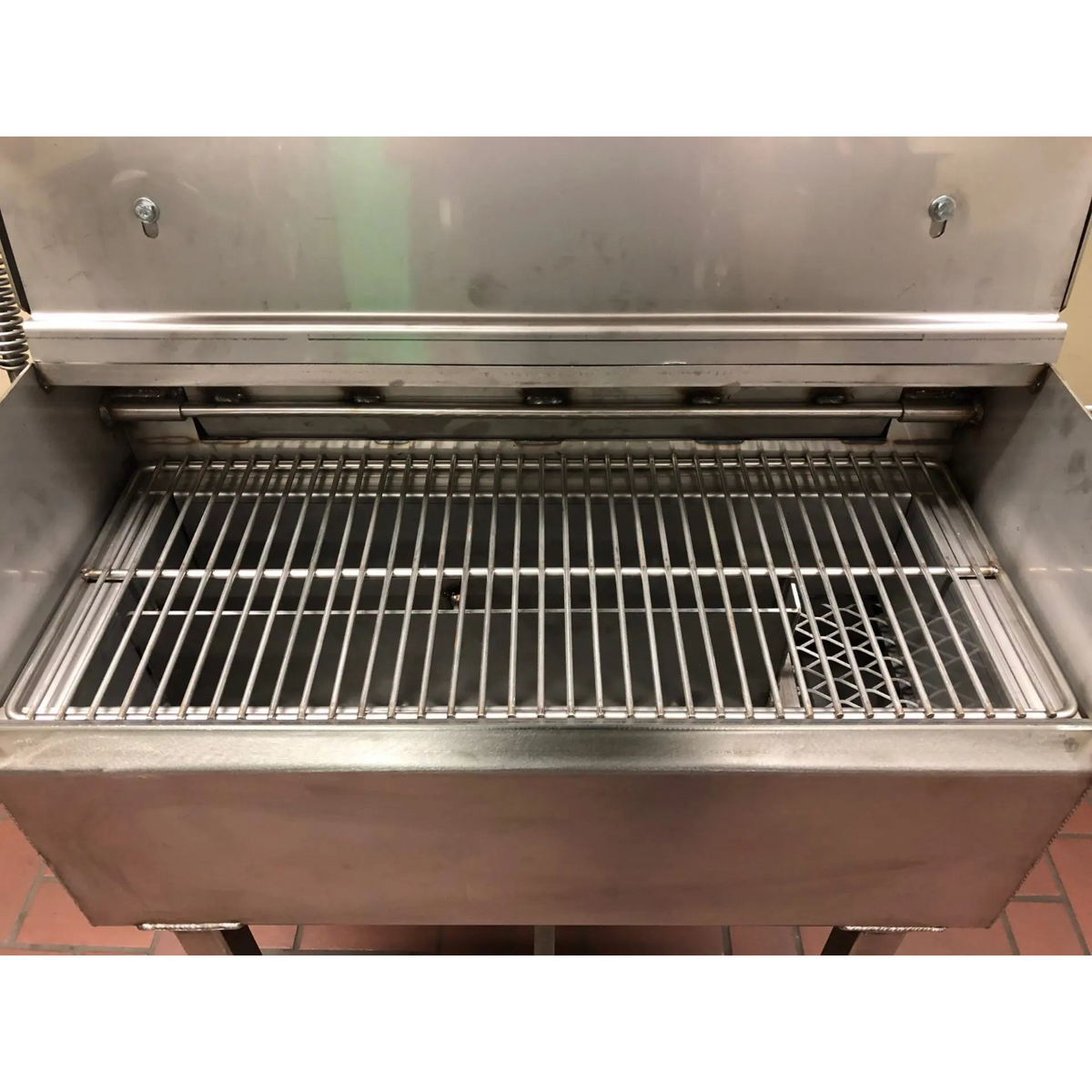 A stainless steel commercial grill or smoker with an open lid and metal grate inside, photographed in a utilitarian setting.