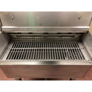 A stainless steel commercial grill or smoker with an open lid and metal grate inside, photographed in a utilitarian setting.