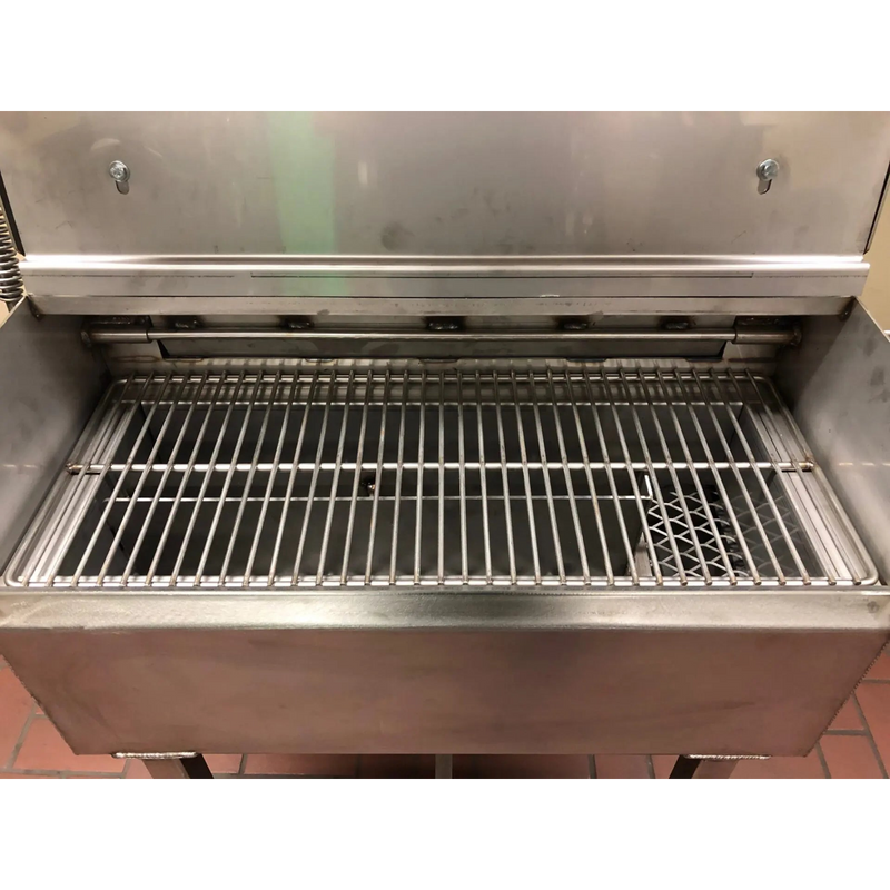 A stainless steel commercial grill or smoker with an open lid and metal grate inside, photographed in a utilitarian setting.
