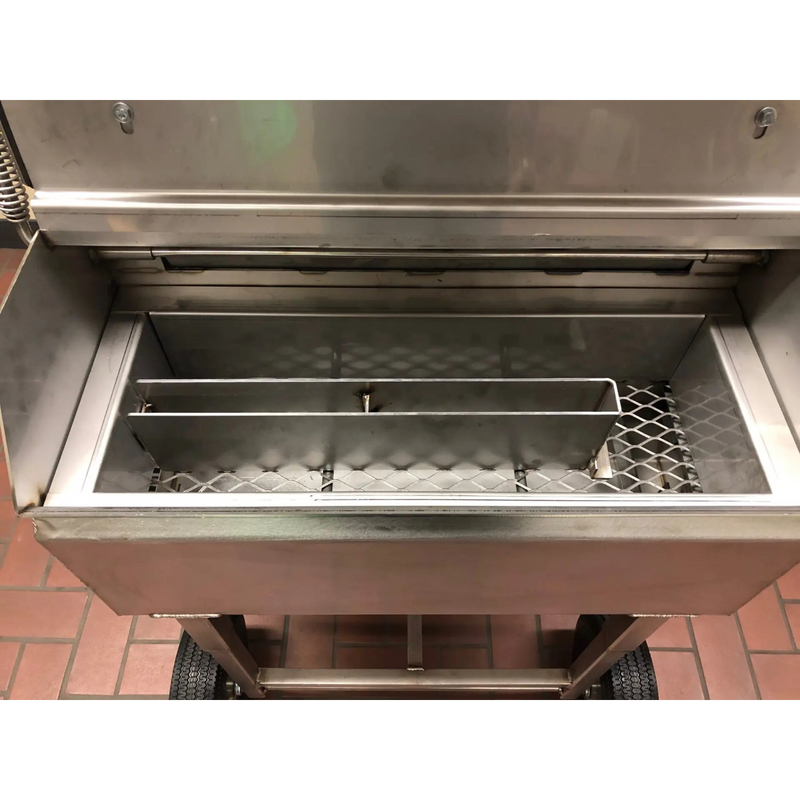 A stainless steel commercial smoker or grill with its lid open, showing the interior chamber, wire racks, and wheels on a tiled floor.
