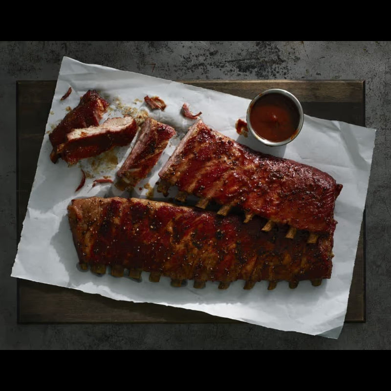 Two racks of glazed barbecue ribs on white parchment paper, served on a dark wooden board with a small metal cup of sauce on a dark textured surface.
