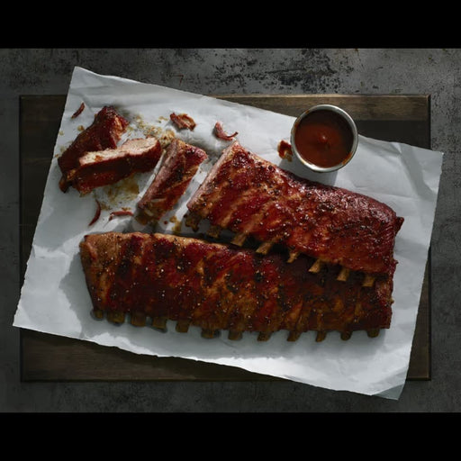Two racks of glazed barbecue ribs on white parchment paper, served on a dark wooden board with a small metal cup of sauce on a dark textured surface.