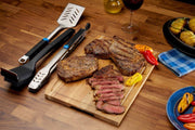 Stainless steel grilling tool set with spatula, tongs, and brush alongside grilled steaks on a wooden cutting board.