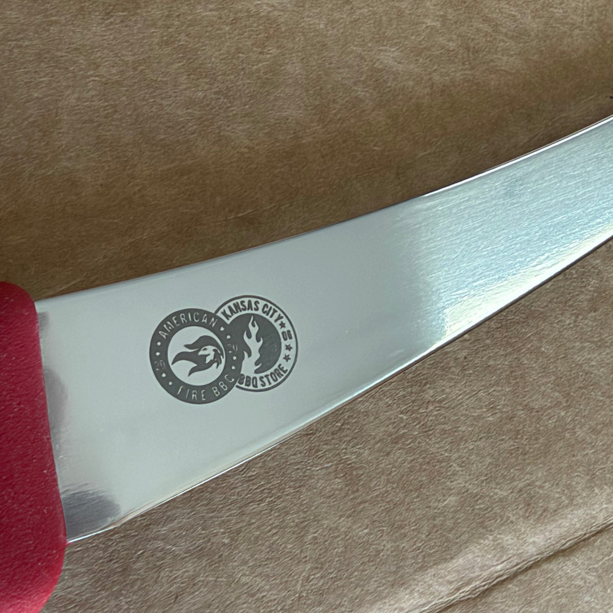 A close-up of a stainless steel knife blade with a circular black logo featuring a horse and rider, resting on a textured brown surface with a red handle visible at the bottom.