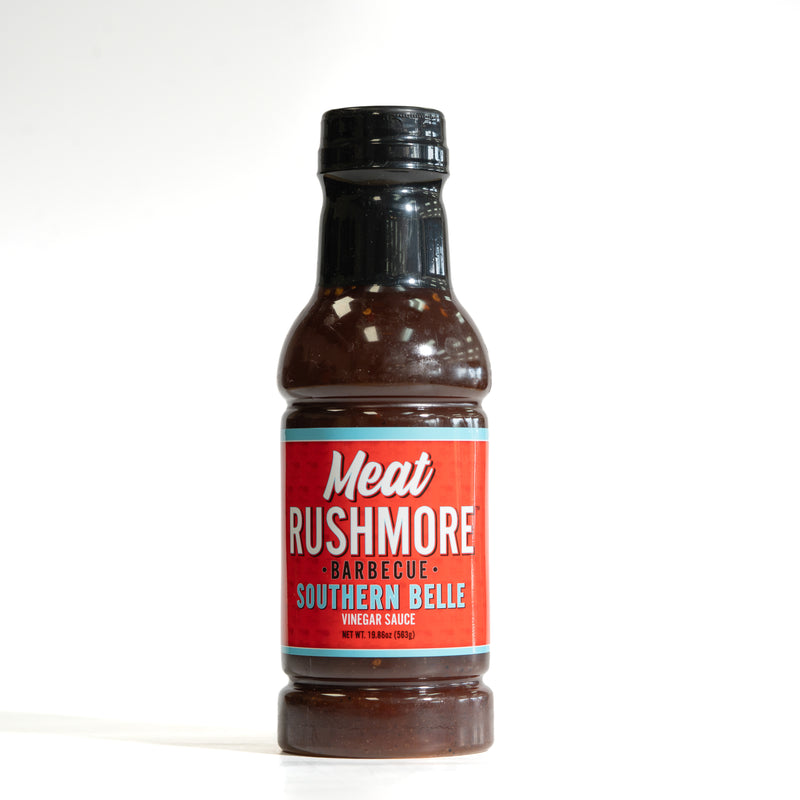 A bottle of Meat Rushmore Barbecue Southern Belle vinegar sauce, featuring a red label with white and blue text, set against a plain white background.