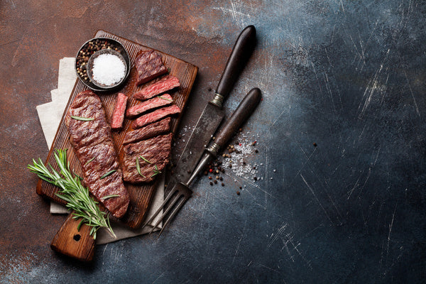 Sliced steak on a wooden cutting board with a knife, salt, and rosemary on a dark surface. The Kansas City BBQ Store