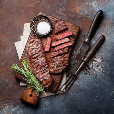 Sliced steak on a wooden cutting board with a knife, salt, and rosemary on a dark surface. The Kansas City BBQ Store